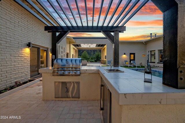 patio terrace at dusk with a sink, an outdoor kitchen, a pergola, and grilling area