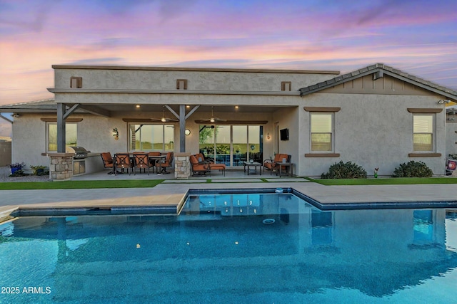 back of property at dusk with ceiling fan, a patio, exterior kitchen, an outdoor pool, and stucco siding