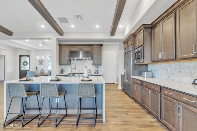 kitchen featuring a breakfast bar area, built in appliances, light wood-type flooring, beamed ceiling, and under cabinet range hood