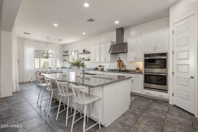 kitchen with appliances with stainless steel finishes, tasteful backsplash, white cabinetry, an island with sink, and wall chimney exhaust hood