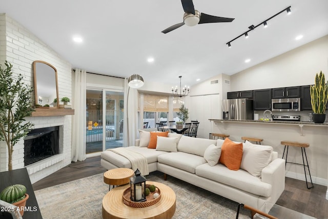 living room featuring lofted ceiling, rail lighting, dark hardwood / wood-style flooring, a brick fireplace, and ceiling fan with notable chandelier