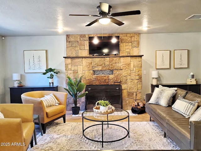 living room featuring ceiling fan, a stone fireplace, a textured ceiling, and light hardwood / wood-style floors