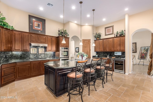 kitchen featuring hanging light fixtures, beverage cooler, a center island with sink, backsplash, and appliances with stainless steel finishes