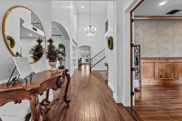 foyer entrance with wallpapered walls, a wainscoted wall, a chandelier, arched walkways, and dark wood finished floors