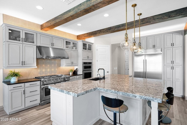 kitchen featuring under cabinet range hood, high quality appliances, a sink, light wood finished floors, and beamed ceiling