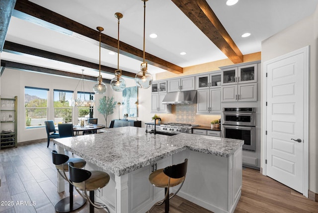 kitchen featuring under cabinet range hood, a sink, stainless steel double oven, beam ceiling, and decorative backsplash