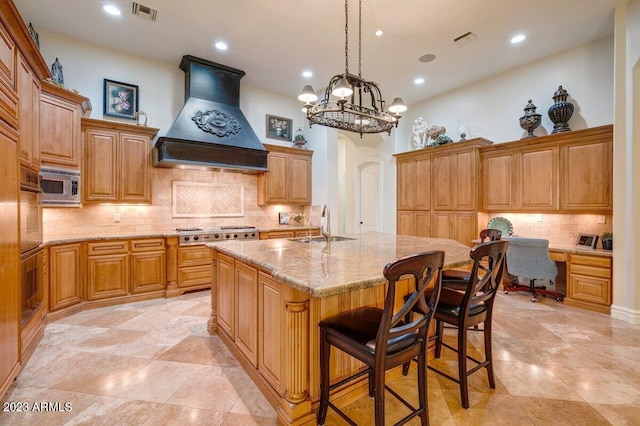 kitchen with appliances with stainless steel finishes, built in desk, sink, custom exhaust hood, and a center island with sink