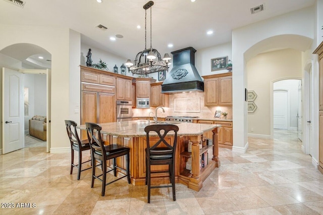 kitchen with premium range hood, tasteful backsplash, built in appliances, light stone counters, and a center island with sink