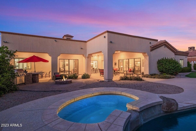 back house at dusk featuring a patio area, an outdoor fire pit, an in ground hot tub, and exterior kitchen