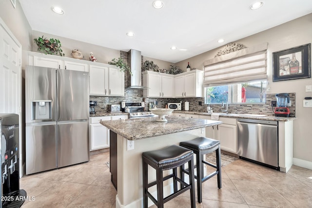 kitchen featuring wall chimney range hood, appliances with stainless steel finishes, light stone counters, white cabinets, and a kitchen island