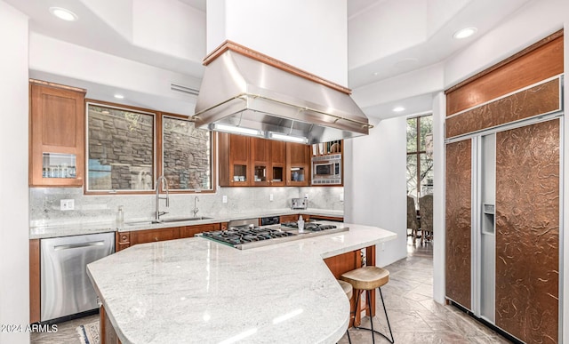 kitchen featuring brown cabinetry, appliances with stainless steel finishes, a sink, island exhaust hood, and backsplash