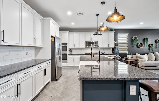 kitchen with appliances with stainless steel finishes, a kitchen island with sink, pendant lighting, dark stone countertops, and light parquet flooring