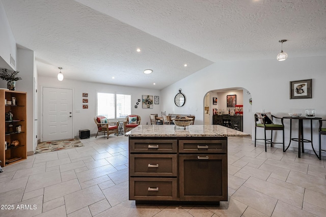 kitchen featuring arched walkways, a center island, vaulted ceiling, dark brown cabinets, and light stone countertops