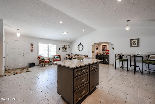 kitchen with light stone counters, arched walkways, a center island, lofted ceiling, and dark brown cabinets