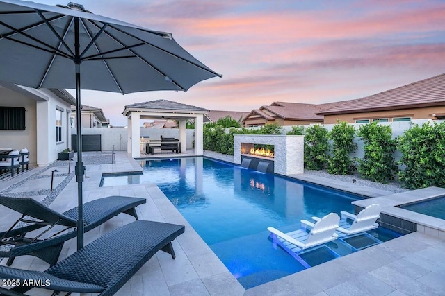 pool at dusk with area for grilling, a warm lit fireplace, a fenced backyard, a gazebo, and a fenced in pool