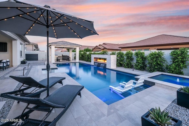 pool at dusk featuring a fenced in pool, a gazebo, a warm lit fireplace, a fenced backyard, and an in ground hot tub