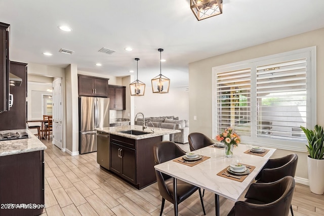 kitchen with a sink, appliances with stainless steel finishes, a center island with sink, dark brown cabinetry, and decorative light fixtures