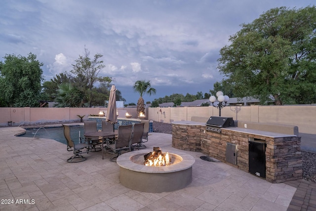 patio terrace at dusk featuring a fire pit, pool water feature, an outdoor kitchen, and grilling area