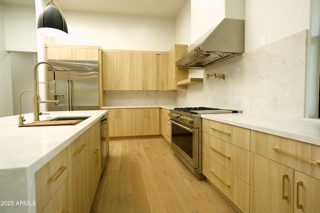 kitchen with premium appliances, hanging light fixtures, light hardwood / wood-style floors, light brown cabinets, and wall chimney exhaust hood