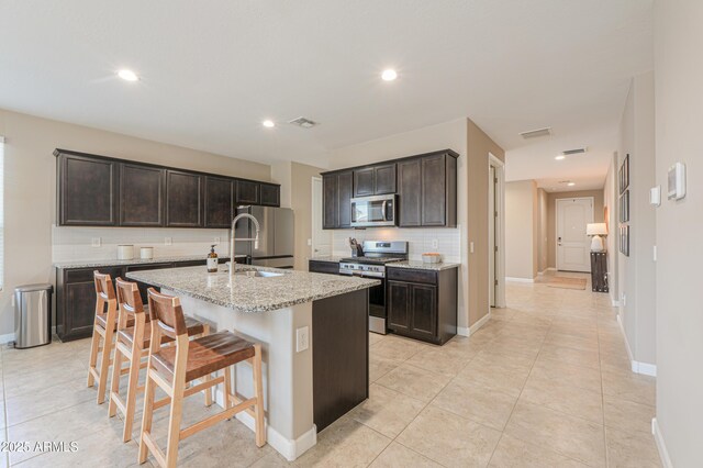 37903 W San Clemente St, Maricopa, AZ 85138 - kitchen featuring stainless steel appliances, decorative backsplash, a center island with sink, light stone countertops, and dark brown cabinets