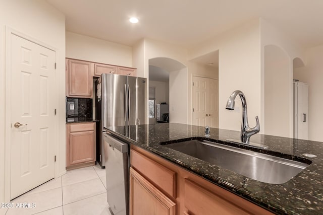 kitchen with light tile patterned floors, arched walkways, a sink, appliances with stainless steel finishes, and dark stone countertops