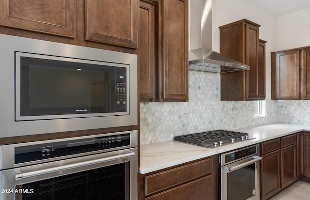 kitchen featuring wall chimney exhaust hood, light stone counters, appliances with stainless steel finishes, and tasteful backsplash