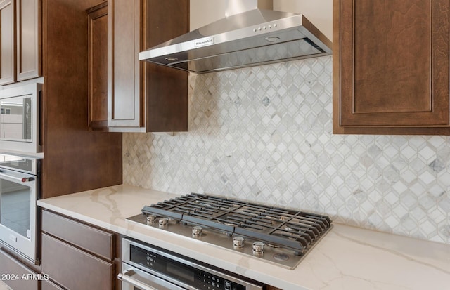 kitchen featuring decorative backsplash, light stone counters, wall chimney range hood, and appliances with stainless steel finishes