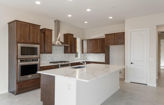 kitchen with sink, stainless steel appliances, wall chimney range hood, light stone counters, and a kitchen island with sink