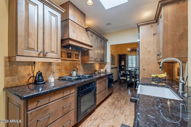 kitchen featuring custom range hood, dark stone counters, black appliances, light hardwood / wood-style floors, and a skylight