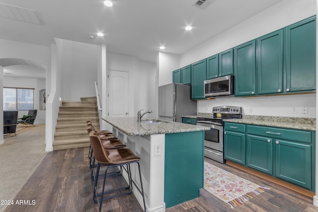 kitchen featuring dark wood-type flooring, sink, an island with sink, stainless steel appliances, and light stone countertops