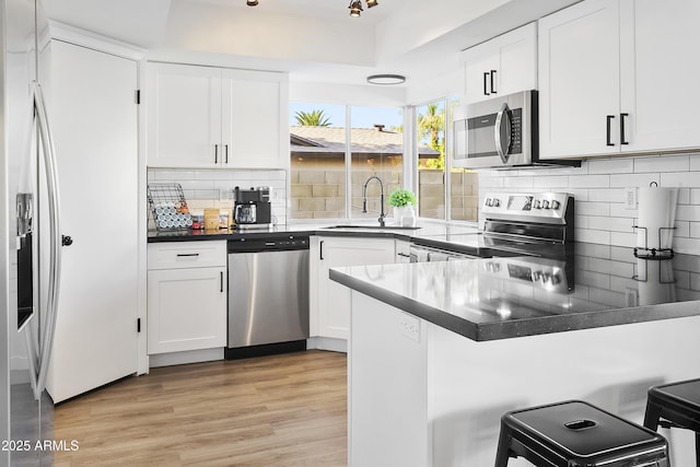 kitchen featuring light wood finished floors, stainless steel appliances, dark countertops, a sink, and a peninsula
