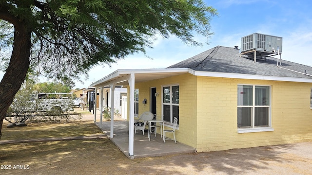 view of side of home featuring roof with shingles, central AC, brick siding, and a patio