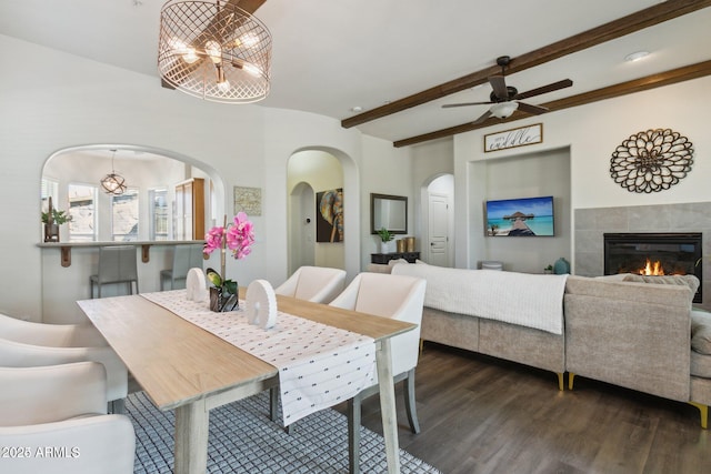 dining room featuring a ceiling fan, arched walkways, dark wood-style flooring, a tiled fireplace, and beamed ceiling