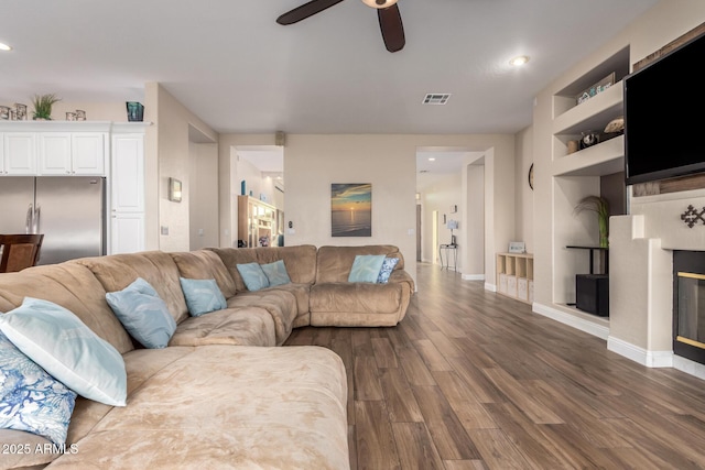 living room with ceiling fan, built in features, baseboards, dark wood-type flooring, and visible vents