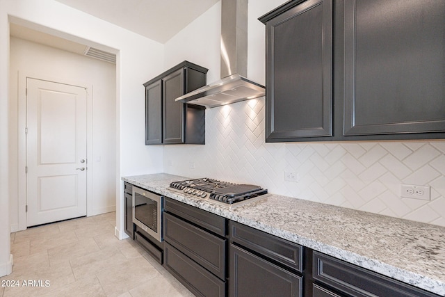 kitchen featuring backsplash, wall chimney range hood, light stone countertops, appliances with stainless steel finishes, and light tile patterned flooring