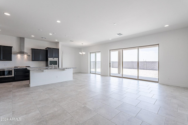 kitchen with stainless steel appliances, wall chimney range hood, light stone counters, backsplash, and an island with sink