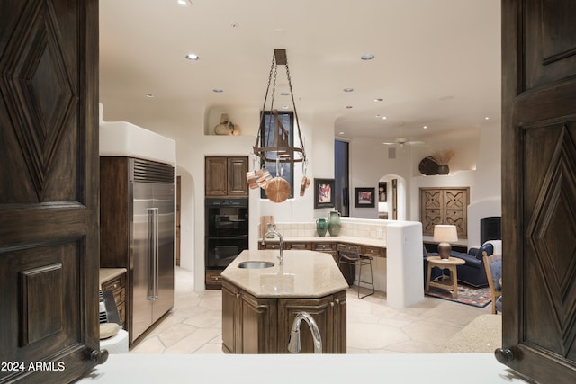 kitchen featuring a center island with sink, built in refrigerator, sink, dark brown cabinetry, and black double oven