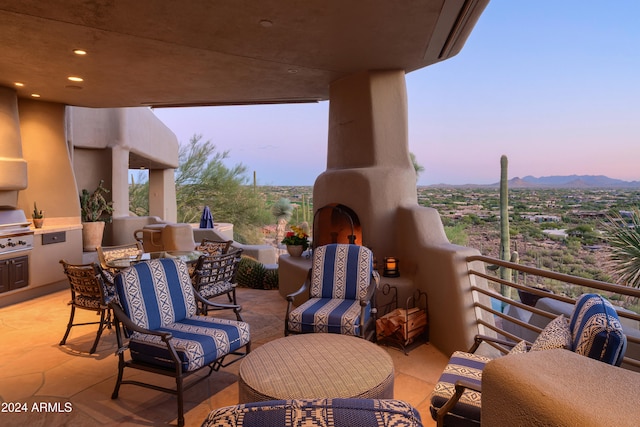 patio terrace at dusk featuring exterior kitchen, area for grilling, an outdoor living space with a fireplace, a mountain view, and a balcony