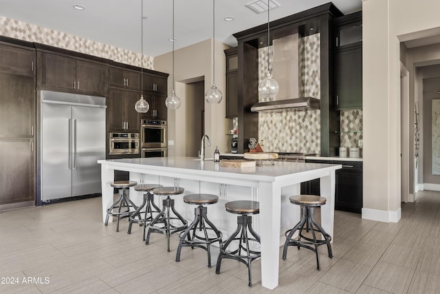 kitchen with wall chimney exhaust hood, stainless steel appliances, backsplash, pendant lighting, and a kitchen island with sink