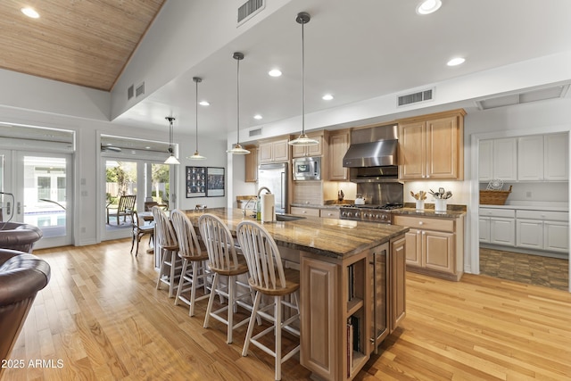 kitchen with dark stone counters, a center island with sink, hanging light fixtures, built in appliances, and wall chimney exhaust hood