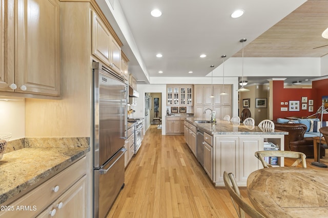 kitchen with light stone counters, high end appliances, sink, light brown cabinets, and hanging light fixtures