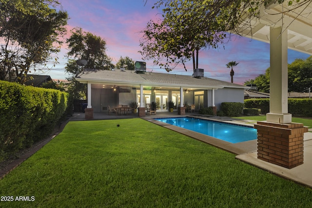 pool at dusk featuring a patio, ceiling fan, and a lawn