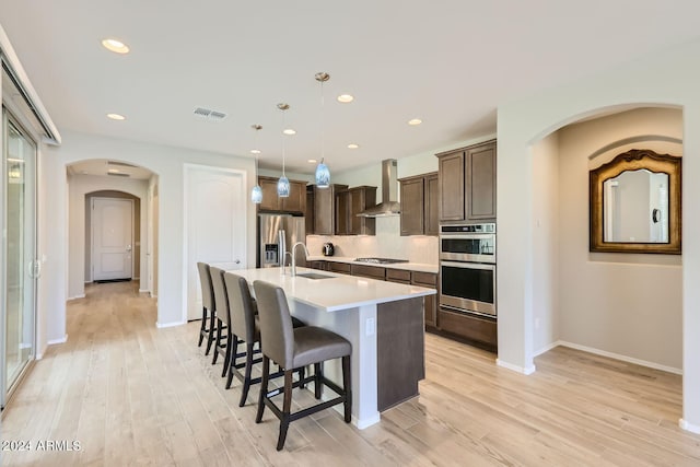 kitchen with a kitchen island with sink, wall chimney range hood, light hardwood / wood-style flooring, appliances with stainless steel finishes, and decorative light fixtures