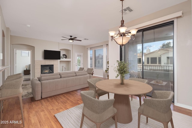 dining space featuring built in shelves, a fireplace, visible vents, light wood-type flooring, and ceiling fan with notable chandelier