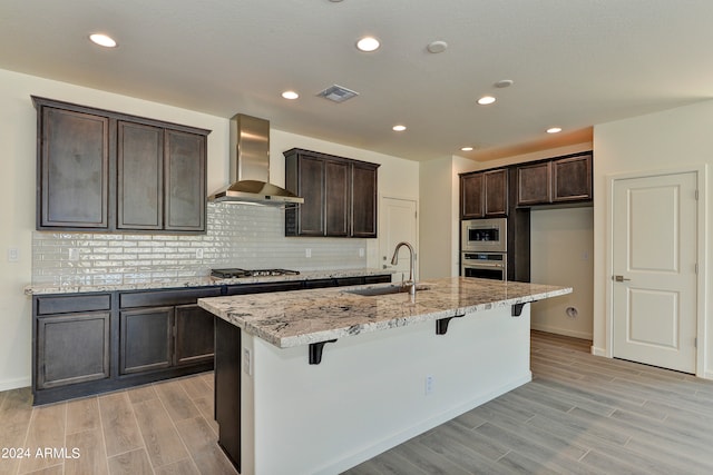 kitchen with light stone counters, sink, a kitchen island with sink, wall chimney range hood, and appliances with stainless steel finishes