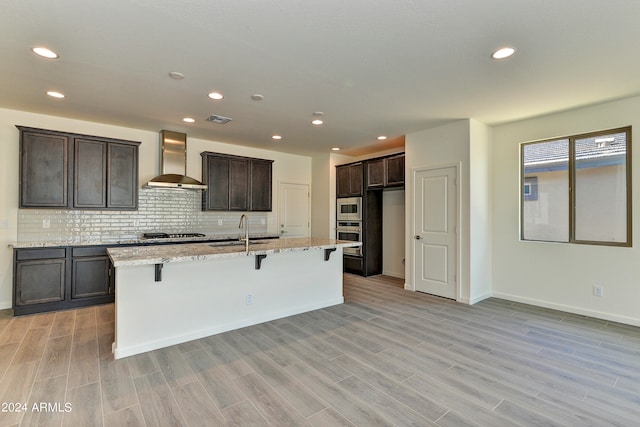 kitchen featuring a center island with sink, stainless steel appliances, wall chimney range hood, and light hardwood / wood-style floors