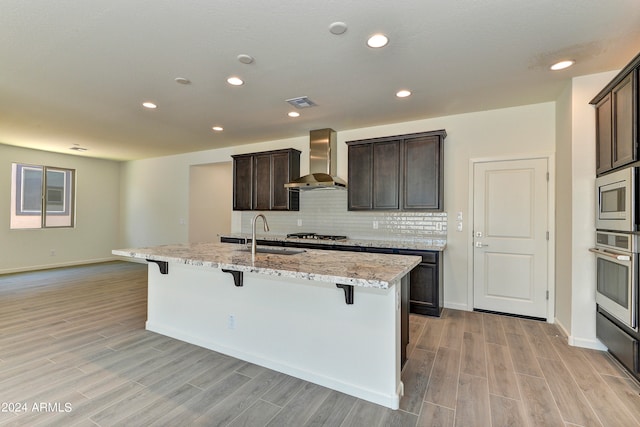 kitchen featuring a kitchen island with sink, light hardwood / wood-style flooring, wall chimney range hood, appliances with stainless steel finishes, and a kitchen bar