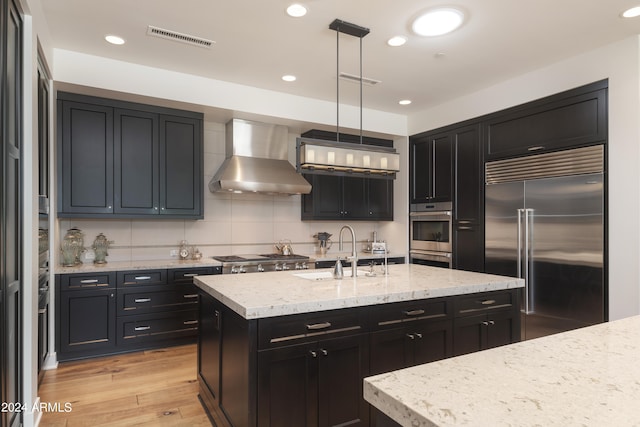 kitchen featuring appliances with stainless steel finishes, wall chimney exhaust hood, sink, tasteful backsplash, and light hardwood / wood-style flooring