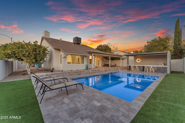 pool at dusk featuring a lawn, a patio, cooling unit, and a bar