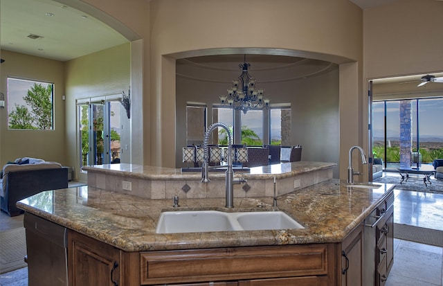 kitchen with ceiling fan with notable chandelier, light stone counters, a kitchen island with sink, and sink